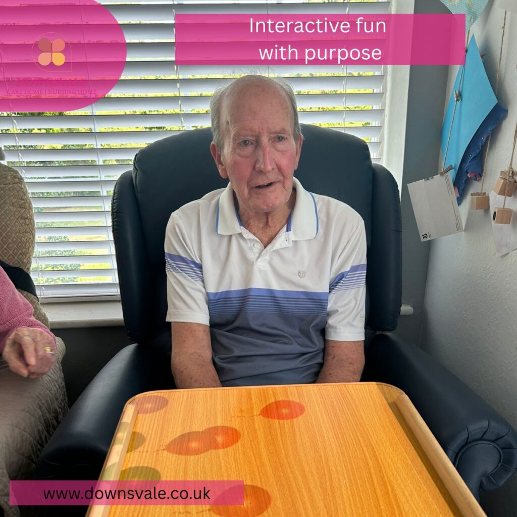 Elderly man seated in a care home chair using an interactive tabletop activity, with colorful visuals on the surface.