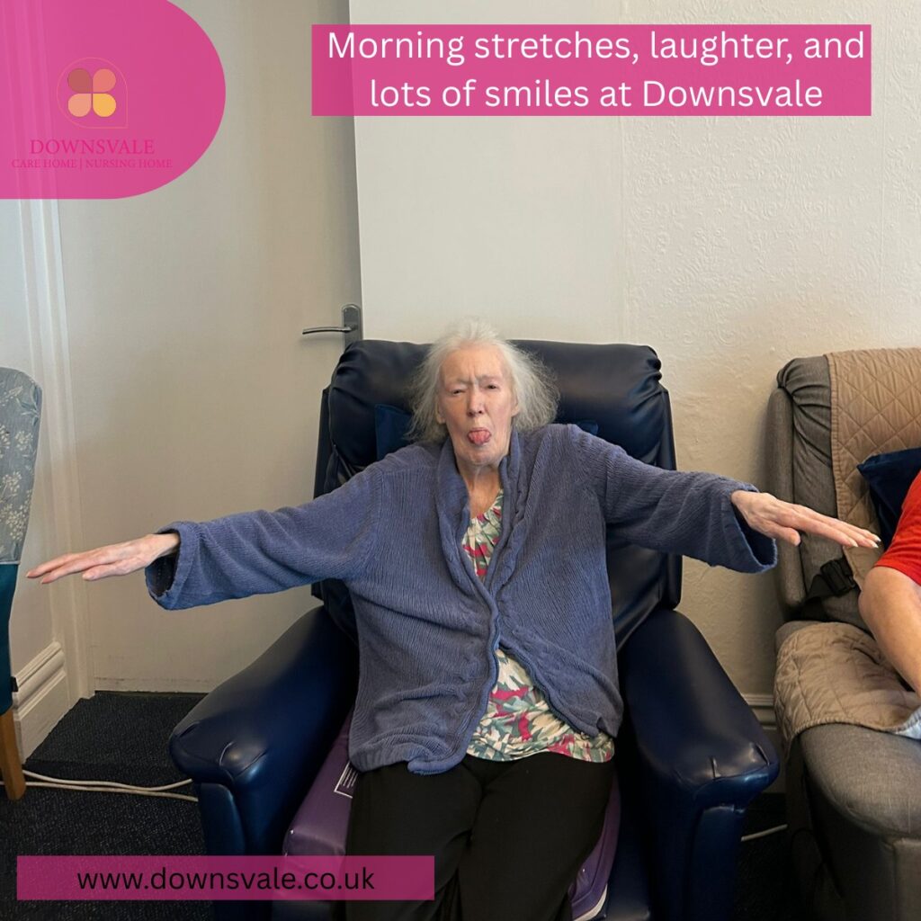 Elderly woman seated in a chair stretching her arms out to the sides during a light exercise session in a care home.