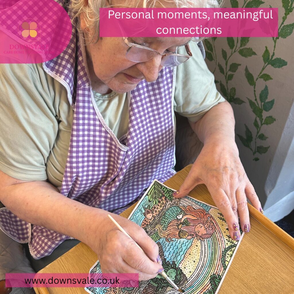 Elderly woman sitting at a table painting a colorful picture, enjoying a creative activity in a care home setting.