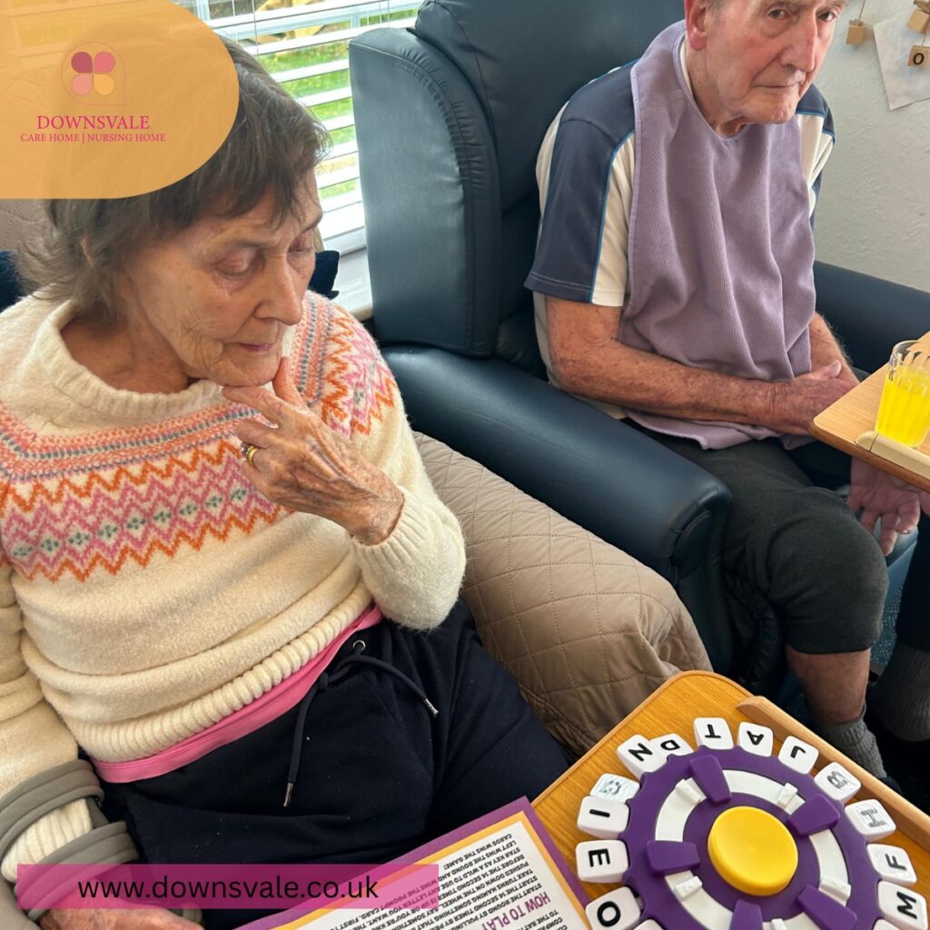 Two elderly residents sitting in armchairs, one engaging with a word-based tabletop game in a care home setting.