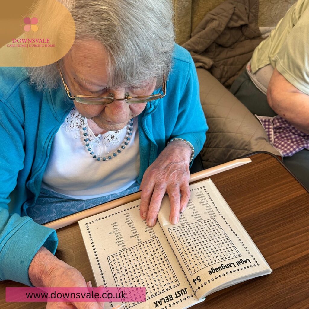 Elderly woman in a care home focusing on a word search puzzle book at a table.