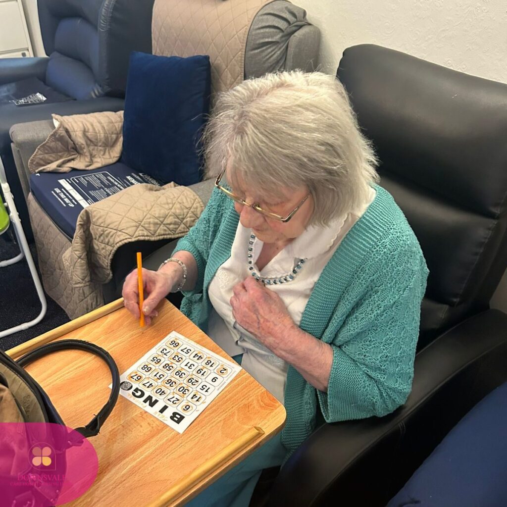 An elderly woman sits at a table marking numbers on a bingo card with a pencil.