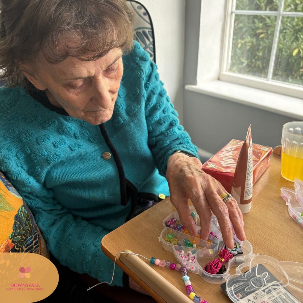 Elderly woman selecting colorful beads from a craft box while making a bracelet at a table.