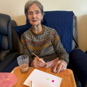 Elderly woman painting a small object while sitting comfortably.