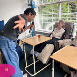 Man singing into a microphone while engaging with an elderly woman seated in a chair by a window.