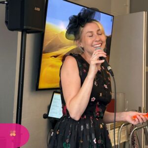 Woman singing into a microphone, smiling, with a speaker and screen behind her.