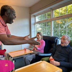 Man interacting animatedly with two elderly residents sitting by a window in a care home.