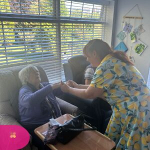 Caregiver holding hands with an elderly woman seated by a window in a care home.