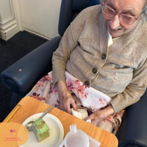 Elderly woman smiling while seated, with a plate of green cake on a table.