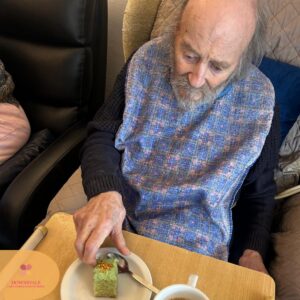 Elderly man sitting in a chair reaching for a small piece of green cake on a plate.