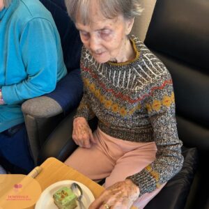 Elderly woman sitting beside another person, looking down at a plate with green cake.