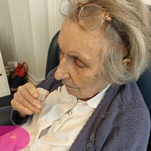 Elderly woman smelling a small bottle while seated in a care home.