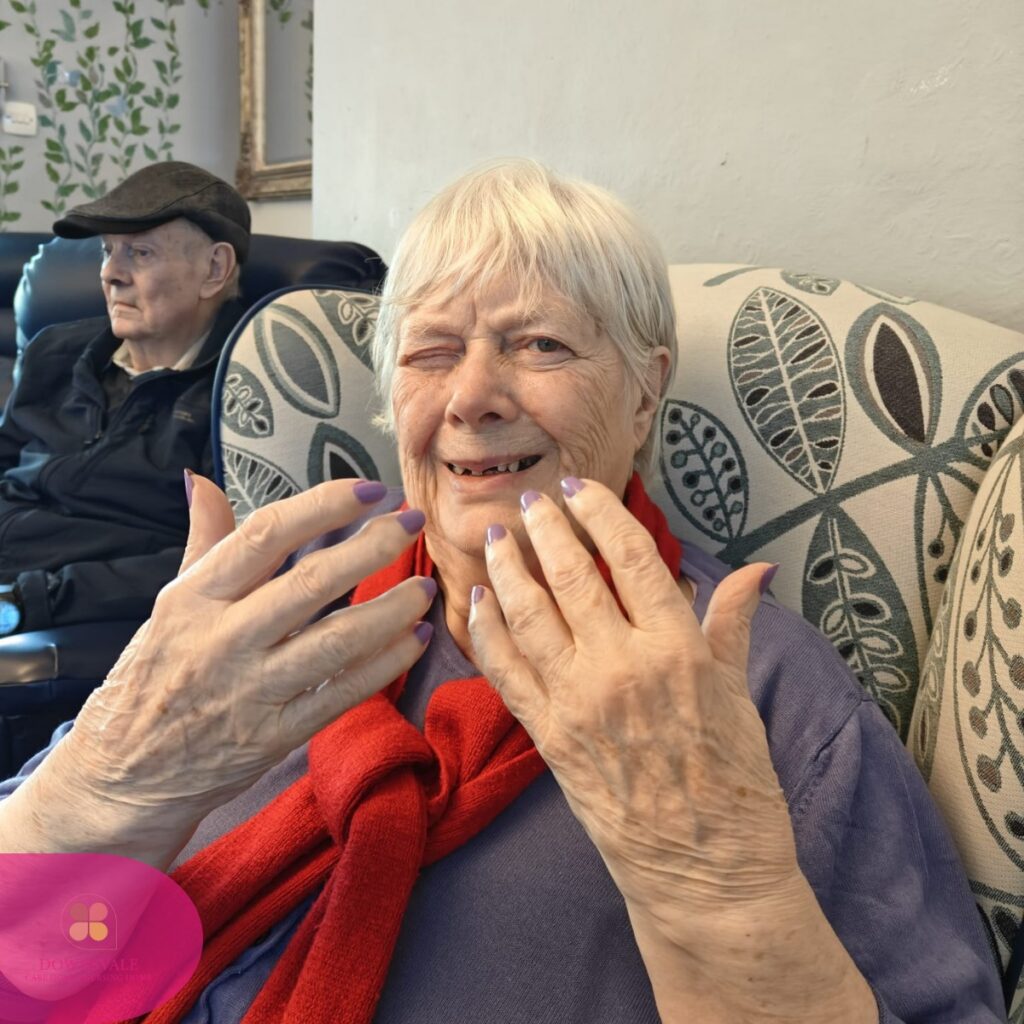 Elderly woman smiling and showing freshly painted nails while seated.