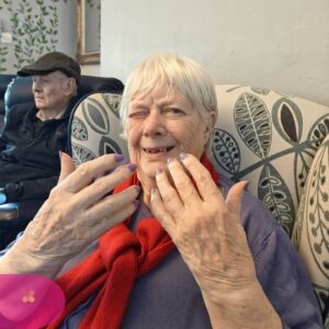 Elderly woman smiling and showing freshly painted nails while seated.