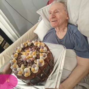 Elderly man in bed smiling at a chocolate cake placed in front of him.