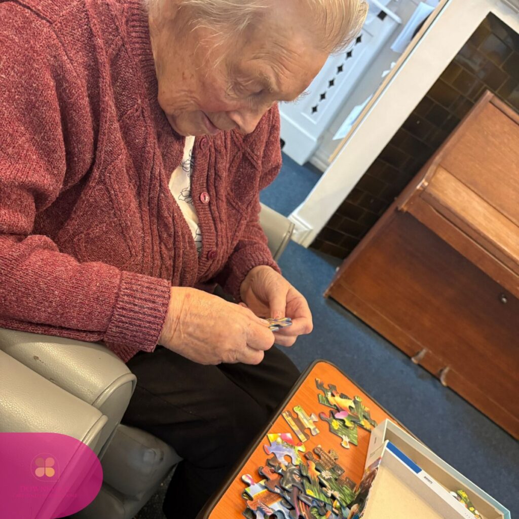 Elderly woman sitting and assembling a jigsaw puzzle at a small table.