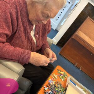 Elderly woman sitting and assembling a jigsaw puzzle at a small table.