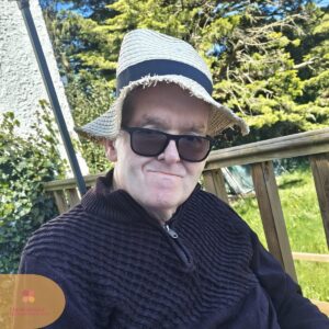 Man wearing sunglasses and a straw hat, sitting outside and smiling slightly.
