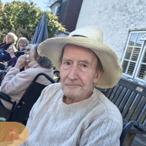 Elderly man wearing a straw hat and sweater, sitting outdoors with others in the background.