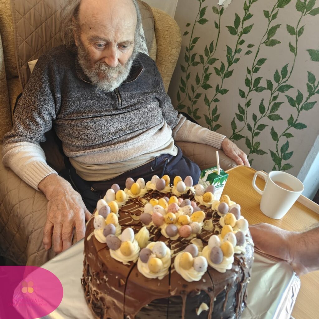 Elderly man sitting in an armchair looking at a chocolate cake on a tray.