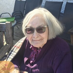 Elderly woman wearing glasses and a purple cardigan, smiling while sitting outdoors in the sunshine.