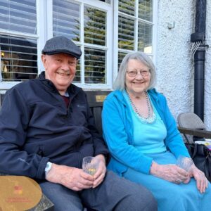 Elderly man and woman sitting on a bench outside, smiling and holding drinks.