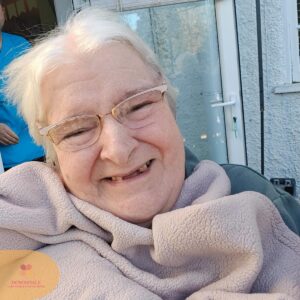 Close-up of an elderly woman smiling, wearing glasses and a cozy fleece jacket.

