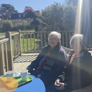 Two older women sitting outdoors at a table with cups, enjoying sunshine on a patio.