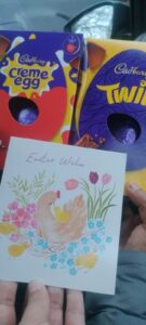 Easter chocolates and a greeting card with a hen and chicks illustration, held in hand.

