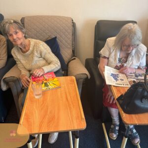 Two elderly women seated with trays, one reading a magazine and the other looking toward the camera.