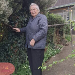 Elderly woman standing in a garden, gently touching a tree branch beside a pathway