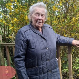 Elderly woman standing by a wooden railing with bright yellow flowers in the background