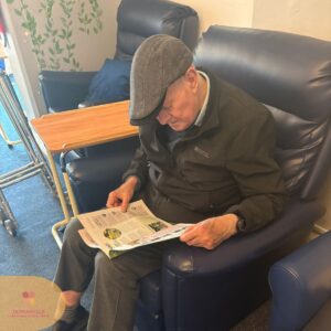 Elderly man sitting in an armchair reading a newspaper.