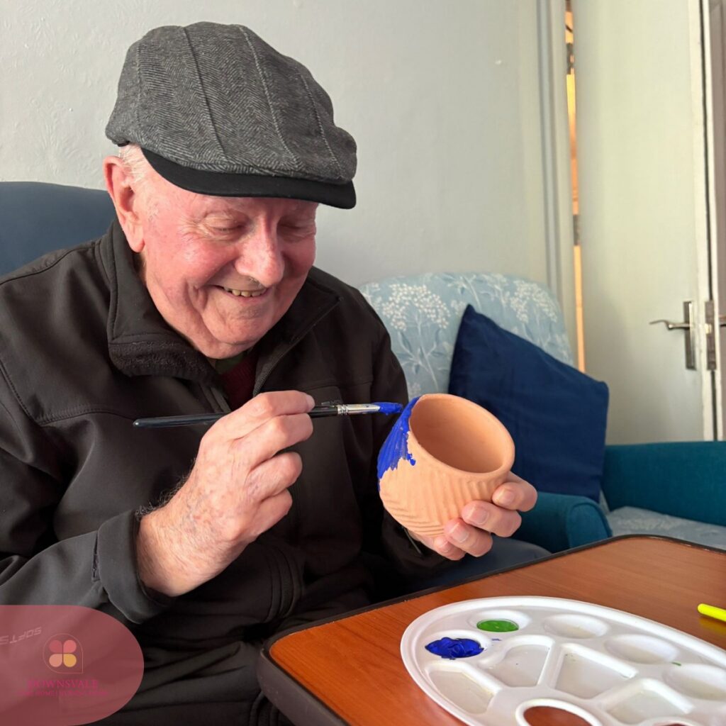 Smiling elderly man painting a small clay pot blue.