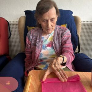 Elderly woman folding pink tissue paper at a table.
