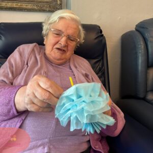Elderly woman shaping a light blue paper flower with a pipe cleaner.

