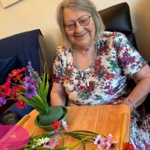Smiling elderly woman seated with a tray, arranging colorful artificial flowers.