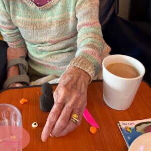 Elderly person crafting with clay at a table beside a cup of tea in a care home.