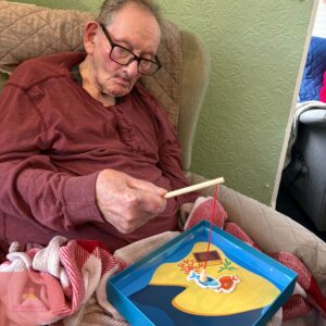 Elderly man enjoying a magnetic fishing game while seated comfortably in a care home chair.