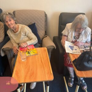Two elderly women sitting comfortably and reading magazines in a care home lounge.