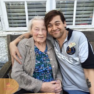 Care staff member and elderly resident smiling together on a garden bench.
