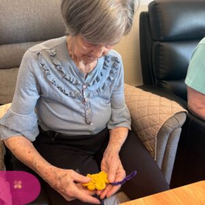 Elderly woman focused on shaping yellow clay during a seated activity.