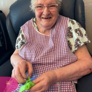 Elderly woman smiling while holding green modeling clay in a chair.