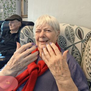Elderly woman smiling and showing freshly painted purple nails in a care home lounge.