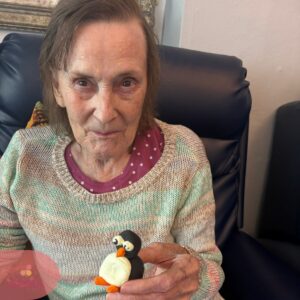 Elderly woman proudly holding a handmade penguin craft and smiling at the camera.