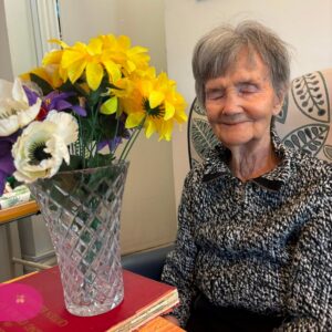 Elderly woman smiling beside a vase of colourful flowers.