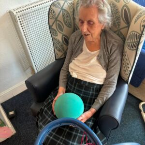 An elderly woman holds a blue ball while seated in an armchair.