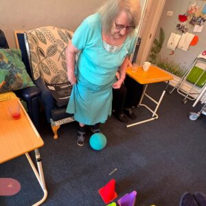 An elderly woman stands beside an armchair with a blue ball at her feet.