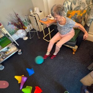 An elderly woman kicks a blue ball while seated in an armchair.