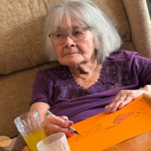 Elderly woman sitting in a chair and drawing on orange paper.